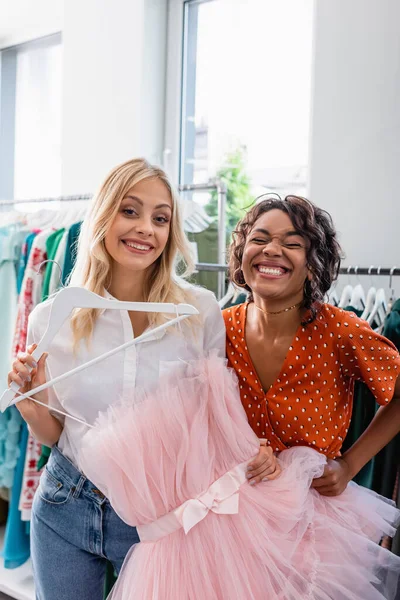 Happy blonde woman holding pink dress near joyful african american friend — Stock Photo