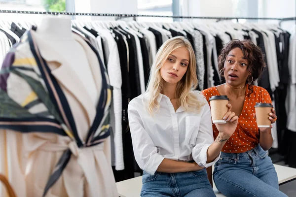 Jovens amigos multiétnicos segurando copos de papel e olhando para roupas borradas na boutique — Fotografia de Stock