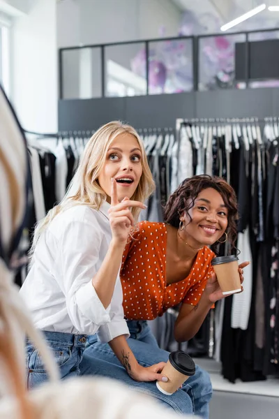 Jeunes amis multiethniques tenant des tasses en papier et pointant du doigt des vêtements en boutique — Photo de stock