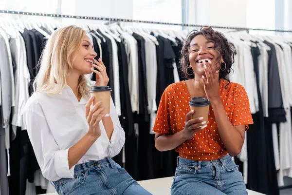 Amigos multiétnicos felices sosteniendo vasos de papel y riendo en boutique - foto de stock