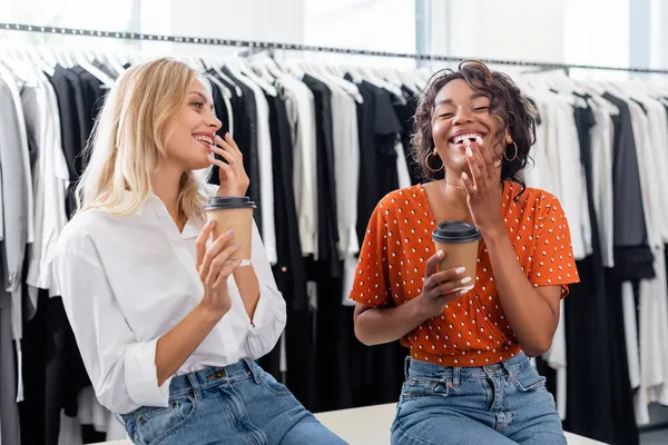 Heureux amis multiethniques tenant des tasses en papier et riant dans la boutique — Photo de stock