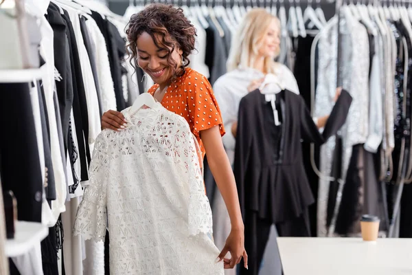 Gioiosa donna africana americana che tiene appendino con abito bianco vicino al cliente offuscata in boutique — Foto stock