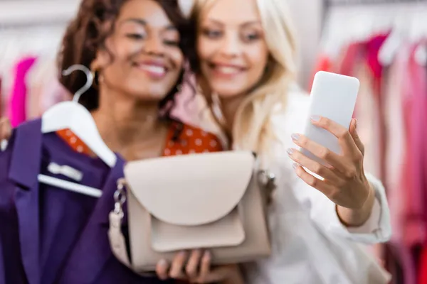 Blurred blonde woman holding purse and taking selfie with cheerful african american friend — Stock Photo