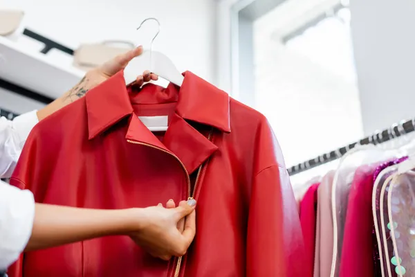 Vista recortada de la mujer tatuada sosteniendo chaqueta de cuero rojo en la percha al elegir la ropa - foto de stock