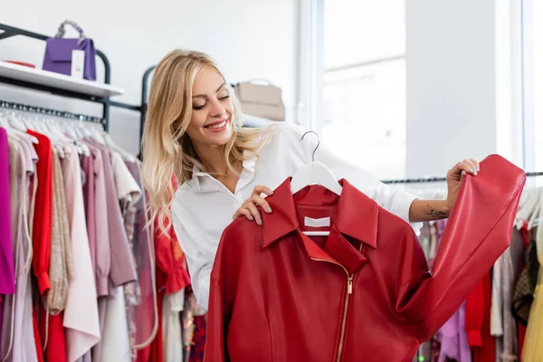 Happy tattooed woman holding red leather jacket on hanger while choosing clothes — Stock Photo