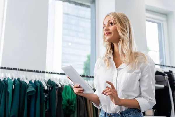 Complacido asistente de ventas en camisa blanca sosteniendo tableta digital en boutique de ropa - foto de stock