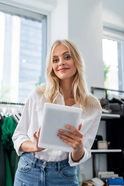 Asistente de ventas sonriente en camisa blanca sosteniendo tableta digital en boutique de ropa - foto de stock