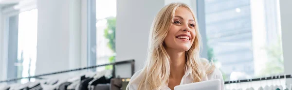 Alegre asistente de ventas en camisa blanca sosteniendo tableta digital en boutique de ropa, bandera - foto de stock