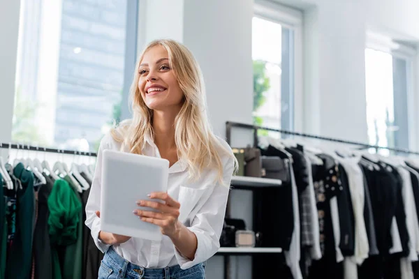 Alegre asistente de ventas en camisa blanca sosteniendo tableta digital en boutique de ropa - foto de stock