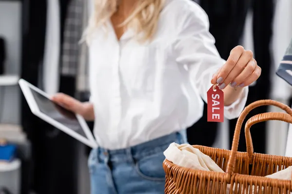 Vue recadrée de l'assistant de vente floue en chemise blanche tenant tablette numérique et étiquette de vente près du panier en osier — Photo de stock