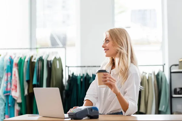 Happy saleswoman holding paper cup near gadgets at sales counter desk — Stock Photo