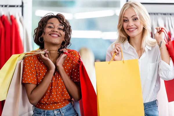 Joyeuses femmes multiethniques tenant des sacs à provisions avec des achats dans la boutique de vêtements — Photo de stock