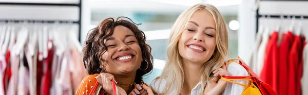 Cheerful interracial women holding shopping bags with purchases in clothing boutique, banner — Stock Photo