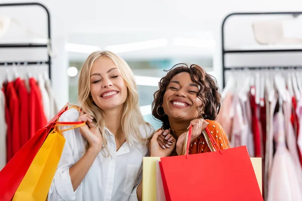 Heureux interracial femmes tenant des sacs à provisions avec des achats dans la boutique de vêtements — Photo de stock