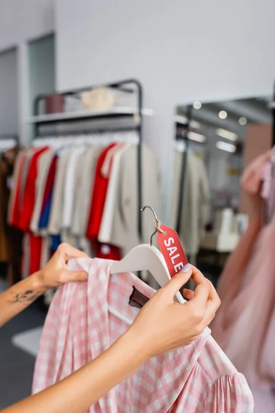 Частичный вид татуированной молодой женщины, держащей вешалку с клетчатым розовым платьем и биркой продажи — стоковое фото