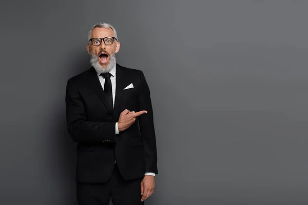 Sorprendido hombre de negocios de mediana edad en gafas que señalan con el dedo en gris - foto de stock
