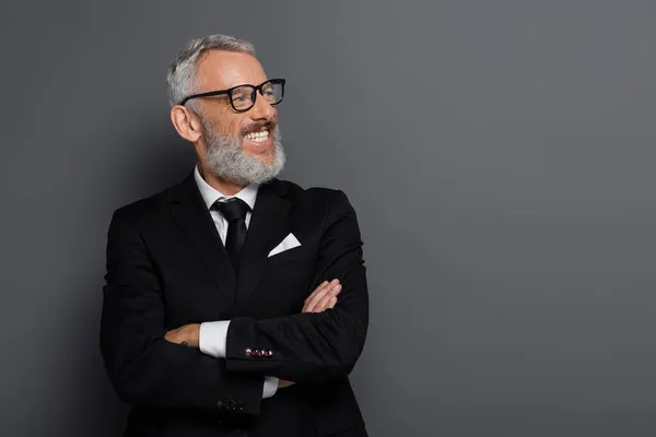 Cheerful middle aged businessman in glasses posing with crossed arms on grey — Stock Photo