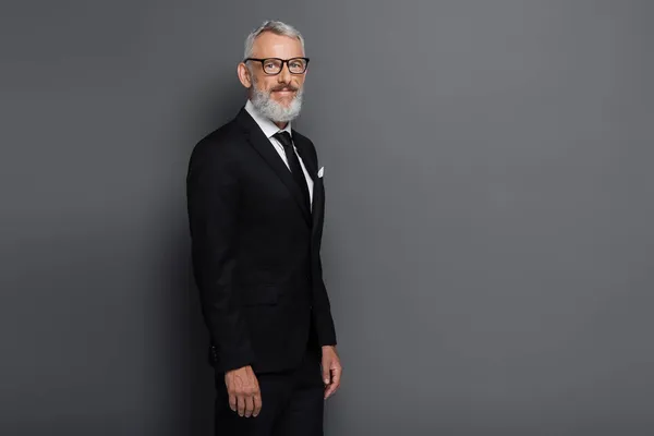 Pleased middle aged businessman in glasses and suit looking at camera isolated on grey — Stock Photo