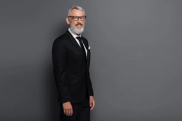 Pleased middle aged businessman in glasses and suit looking at camera isolated on grey — Stock Photo