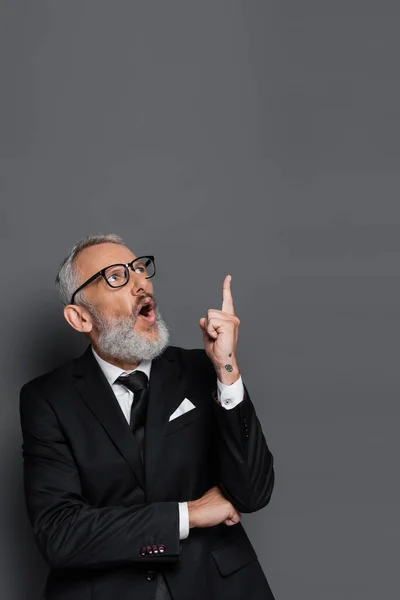 Überraschter Geschäftsmann mittleren Alters in Brille zeigt mit erhobenem Zeigefinger auf Grau — Stockfoto