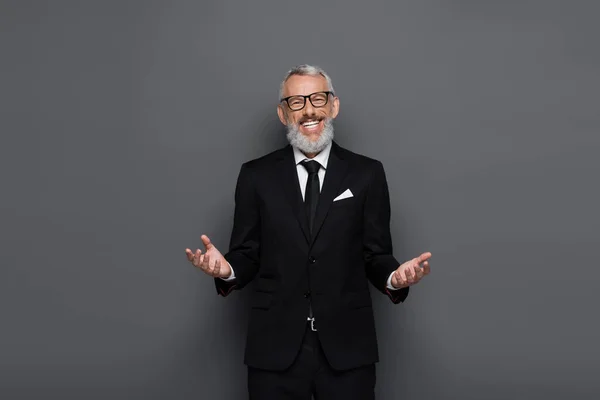 Complacido hombre de negocios de mediana edad en gafas y traje mirando a la cámara con gesto acogedor aislado en gris - foto de stock