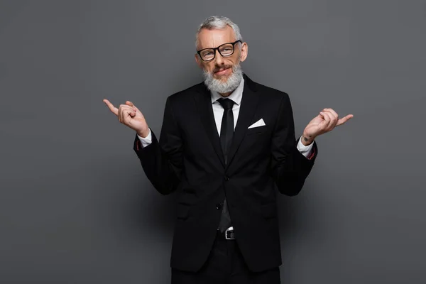Hombre de negocios barbudo de mediana edad en gafas apuntando con los dedos a un lado en gris - foto de stock