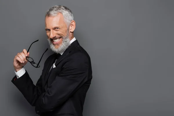 Homem de negócios de meia idade alegre segurando óculos e sorrindo em cinza — Fotografia de Stock