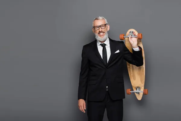 Joyful middle aged businessman in suit and glasses holding longboard on grey — Stock Photo