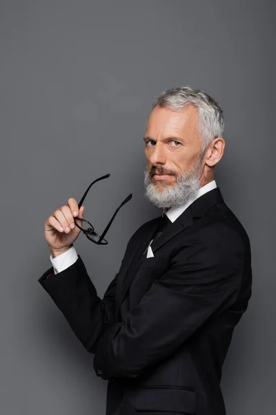 Middle aged businessman in suit holding glasses on grey — Stock Photo