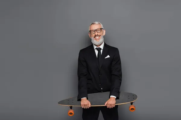 Happy middle aged businessman in suit and glasses holding longboard on grey — Stock Photo