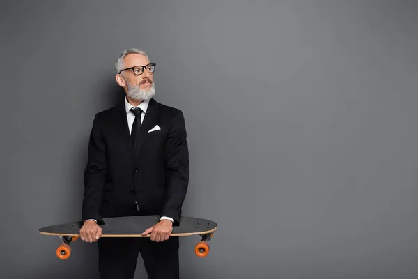 Homme d'affaires barbu d'âge moyen en costume et lunettes tenant longboard sur gris — Photo de stock