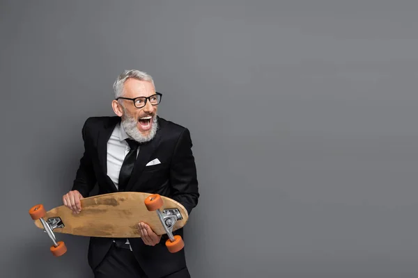 Middle aged businessman in suit and glasses holding longboard and screaming on grey — Stock Photo