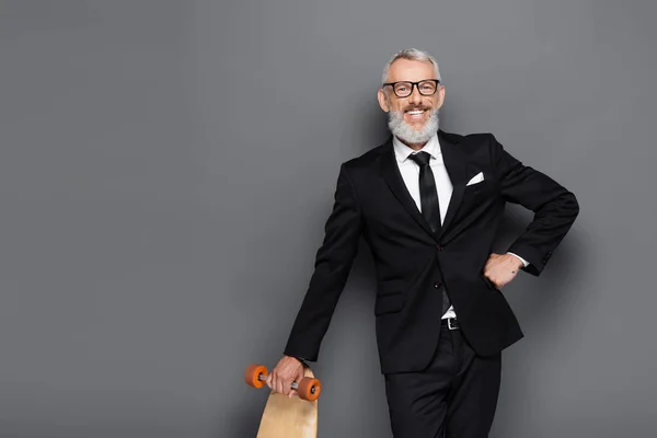 Happy middle aged businessman in suit and glasses holding longboard while posing with hand on hip on grey — Stock Photo