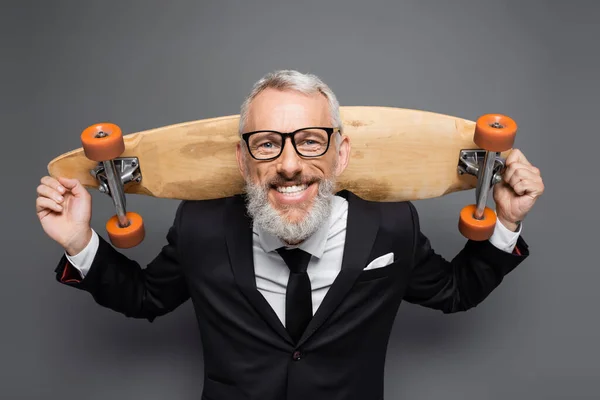 Positive middle aged businessman in suit and glasses holding longboard on grey — Stock Photo