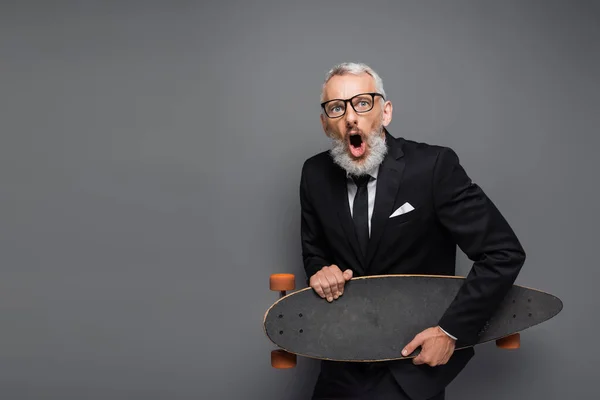 Shocked middle aged businessman in suit and glasses holding longboard isolated on grey — Stock Photo