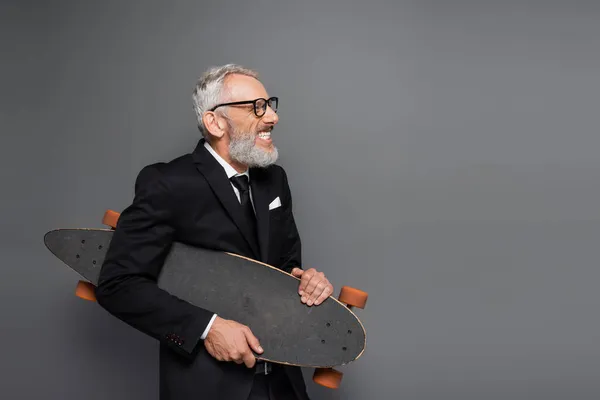 Joyful middle aged businessman in suit and glasses holding longboard on grey — Stock Photo