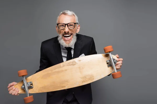 Joyful and mature businessman in suit and glasses holding longboard isolated on grey — Stock Photo