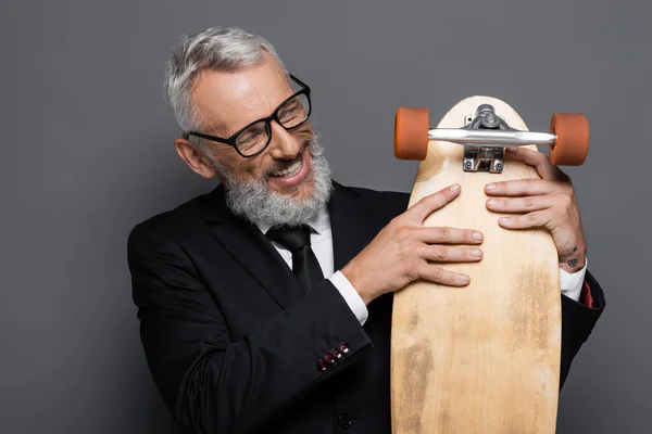 Happy and mature businessman in glasses holding longboard on grey — Stock Photo