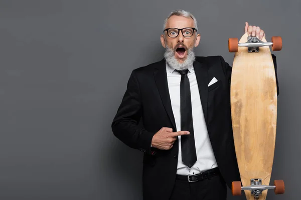 Choqué homme d'affaires d'âge moyen en costume et lunettes pointant vers longboard sur gris — Photo de stock