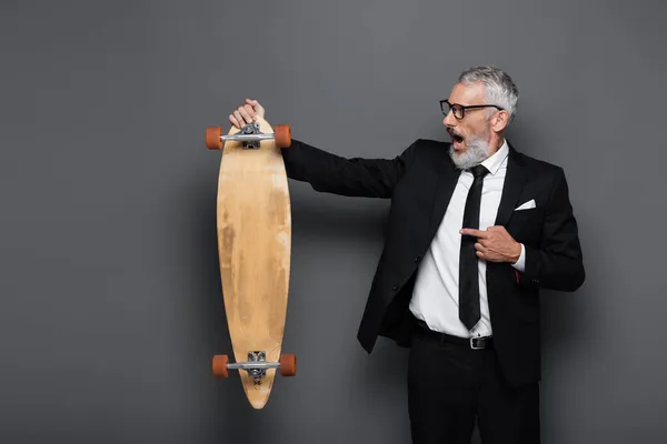 Shocked and mature businessman in suit and glasses pointing at longboard on grey — Stock Photo