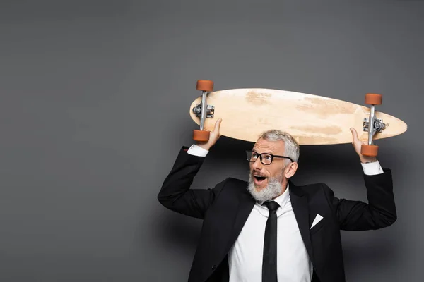 Amazed and mature businessman in suit and glasses holding longboard above head on grey — Stock Photo