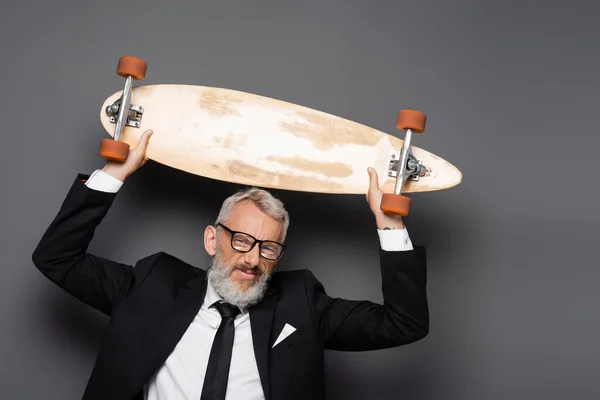 Joyful and mature businessman in suit and glasses holding longboard above head on grey — Stock Photo