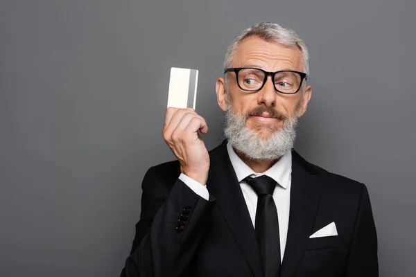Радісний і зрілий бізнесмен в костюмі і окулярах тримає кредитну картку ізольовано на сірому — стокове фото