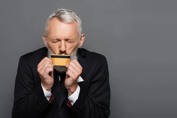 Mature businessman in suit kissing credit card isolated on grey — Stock Photo