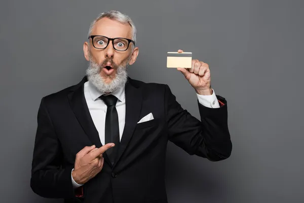 Shocked and mature businessman in suit and glasses pointing at credit card isolated on grey — Stock Photo