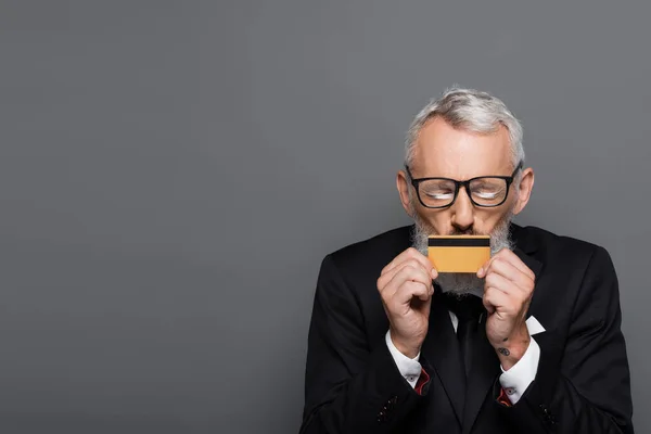 Mature businessman in glasses kissing credit card isolated on grey — Stock Photo