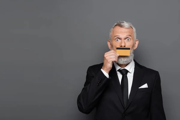 Homme d'affaires mature en costume couvrant la bouche avec carte de crédit isolé sur gris — Photo de stock