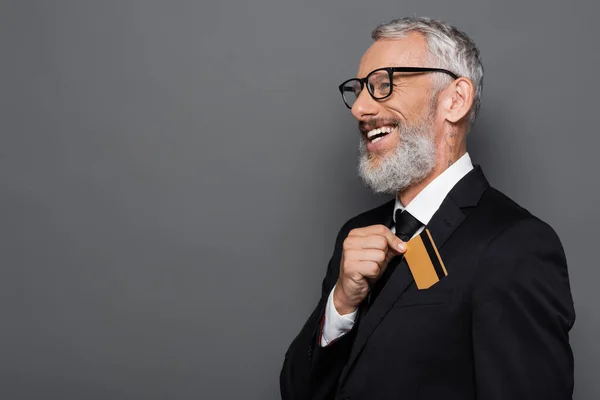 Щасливий середнього віку бізнесмен в костюмі і окулярах кладе кредитну картку в кишеню ізольовано на сірому — стокове фото