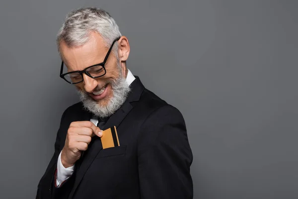 Alegre hombre de negocios de mediana edad en traje y gafas de poner la tarjeta de crédito en el bolsillo aislado en gris - foto de stock