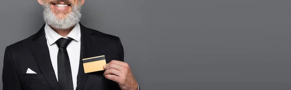 Vue recadrée de joyeux homme d'affaires d'âge moyen en costume tenant la carte de crédit isolée sur gris, bannière — Photo de stock